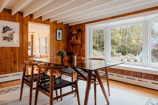 Photograph of a lake house dining room overlooking a green forest outside, the indoors decorated with lake house wall art and a dining set