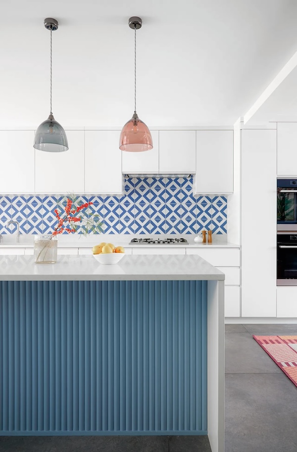 Photograph of a kitchen with a reeded wood kitchen island in the foreground and white cabinetry and a blue and white tile backsplash in the background