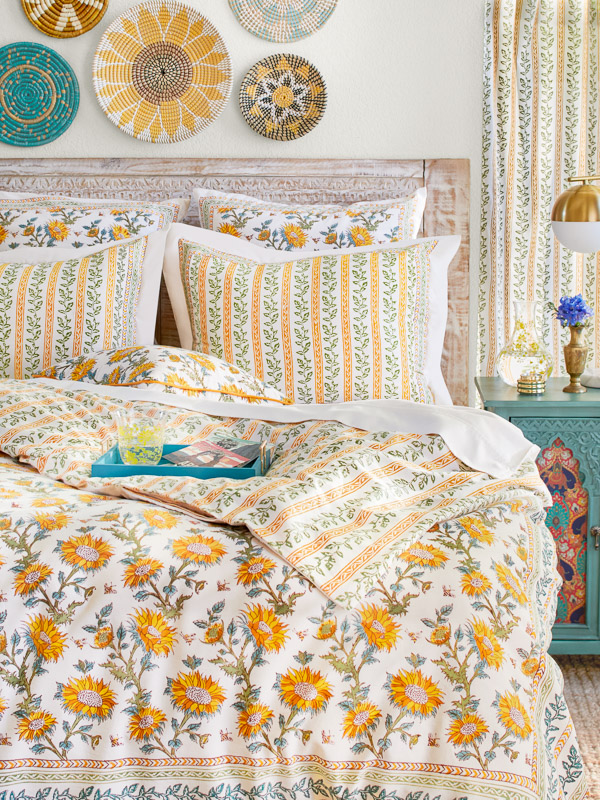 Photograph of a bedroom decorated with sunflower fabric bedding and bright, organic accents such as radial wall baskets, coordinating botanic stripe curtains, and a floral water carafe.